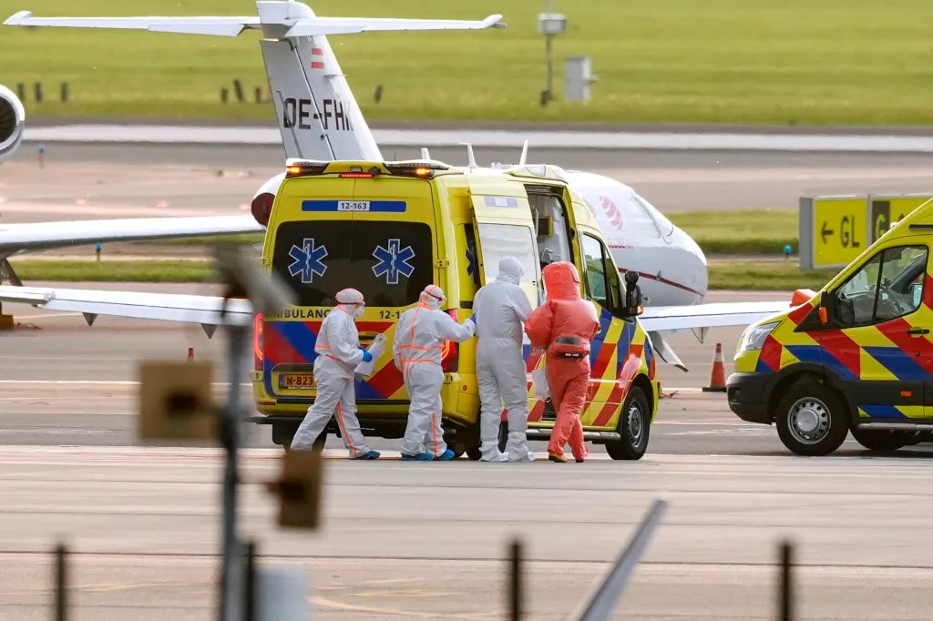 Medics in protective gear escorting a patient in a red suit into an ambulance at an airport.