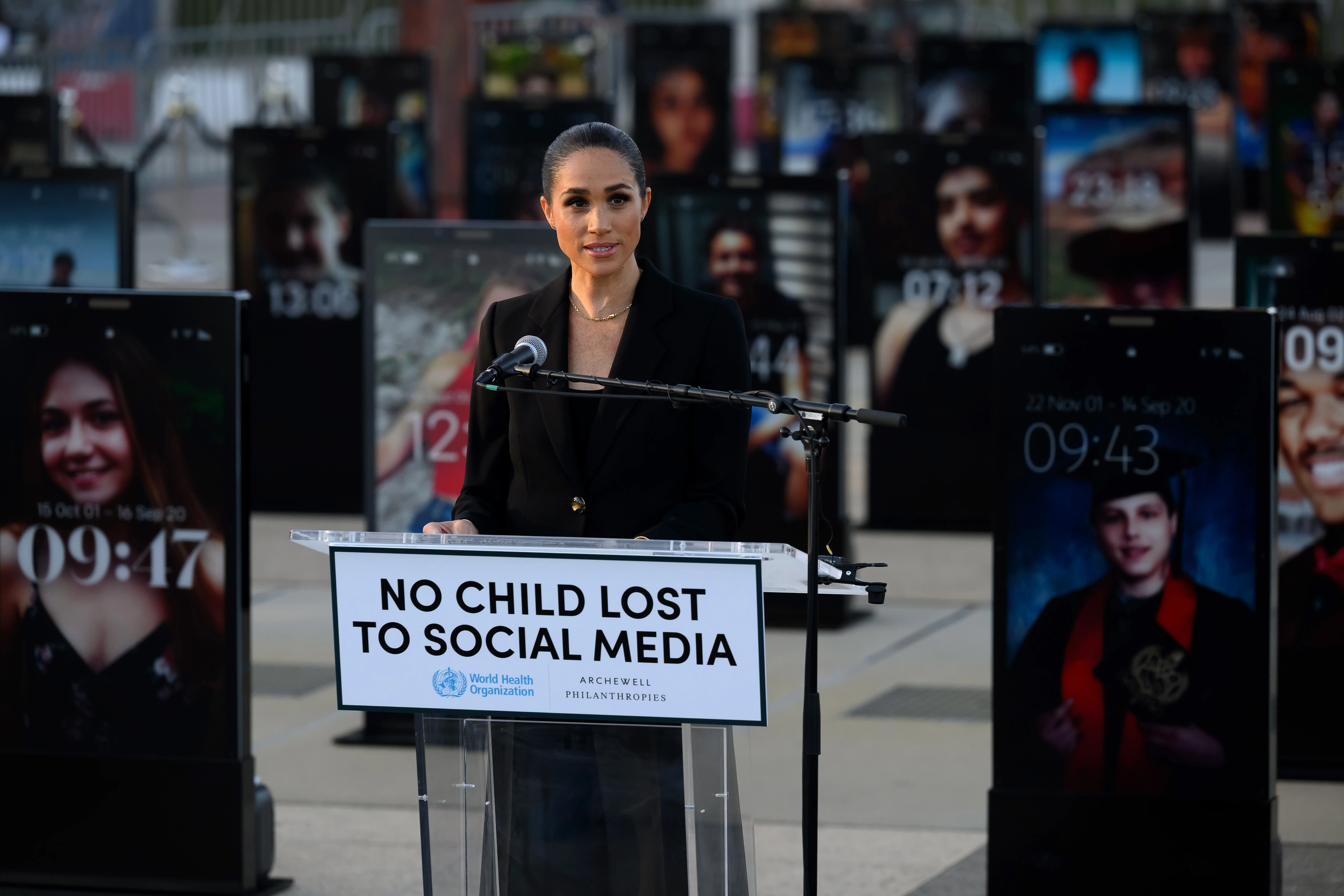 Meghan Markle speaking at a podium that says "NO CHILD LOST TO SOCIAL MEDIA" in front of an installation of illuminated smartphone lock screens.