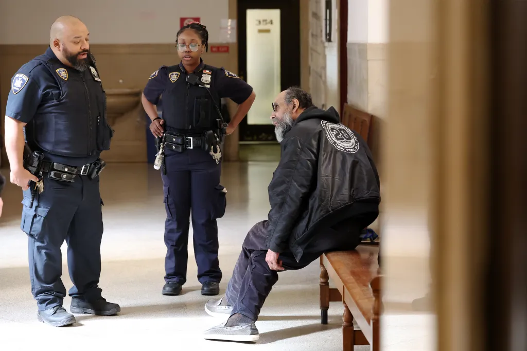 Two police officers stand in a courthouse hallway next to Melvin Dejesus, who sits on a bench.