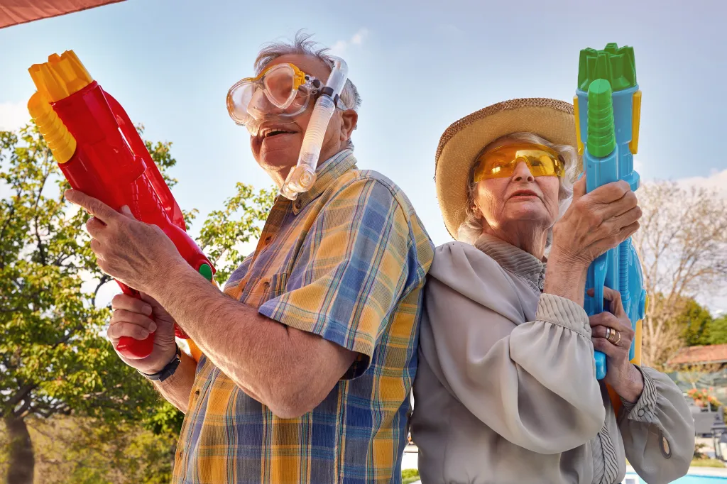 A senior man in a snorkel mask and a senior woman in a straw hat and yellow glasses standing back-to-back, each holding a water gun.