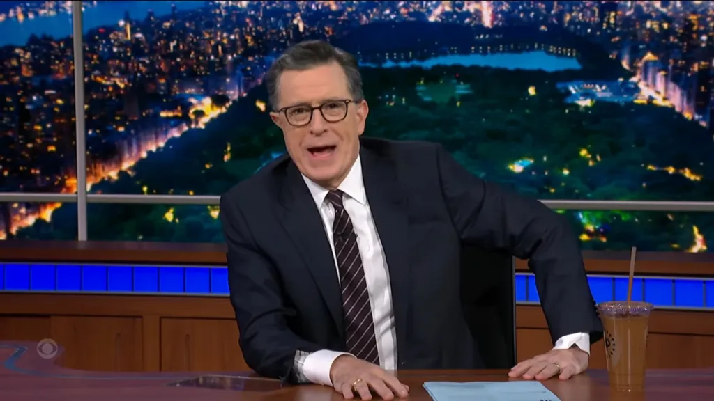 Stephen Colbert hosting "The Late Show" from his desk with a cityscape backdrop.