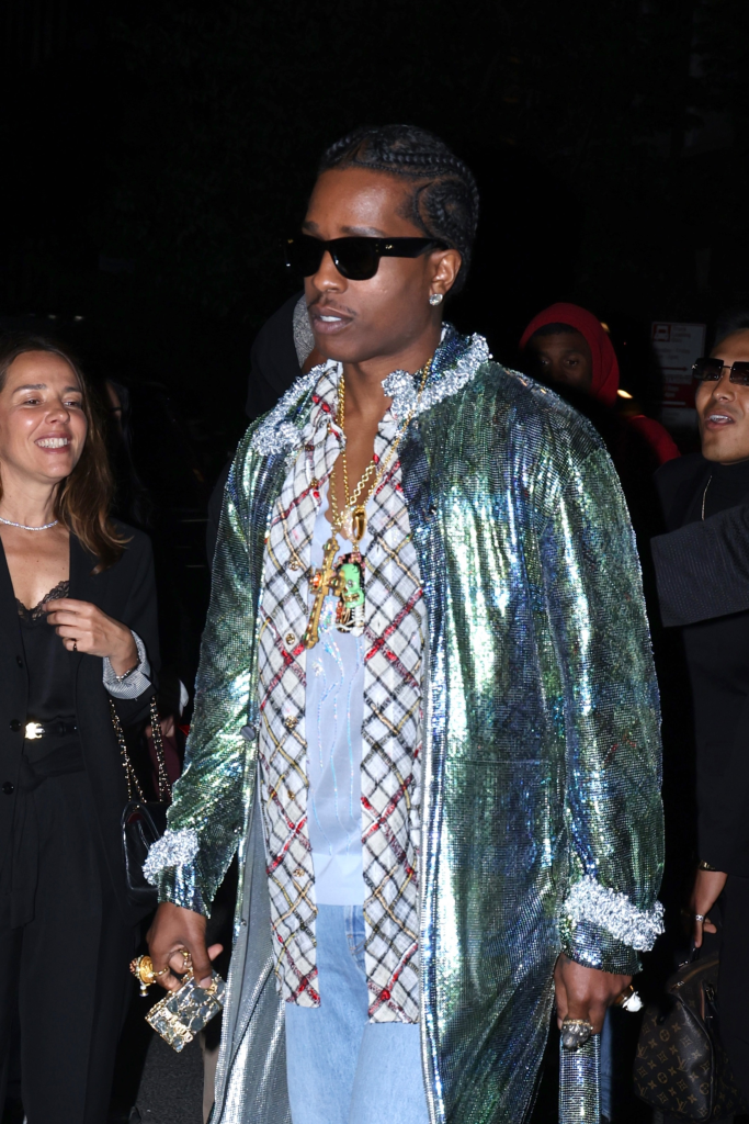 A$AP Rocky wearing a metallic green and blue sequined coat over a plaid and light blue shirt, light wash jeans, and dark sunglasses.