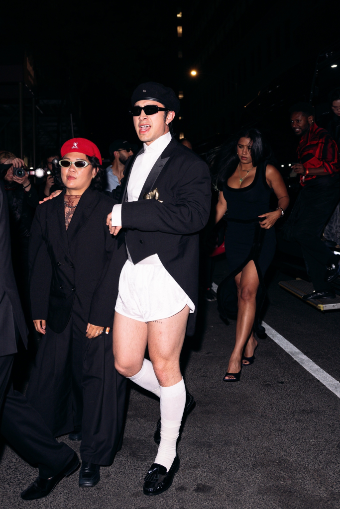 Hudson Williams wearing a tuxedo blazer, white shorts, knee-high socks, dress shoes, black sunglasses, and a black hat, arriving at the GQ after-party in London.