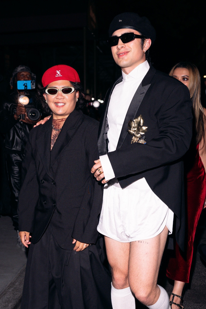 Hudson Williams in a black tuxedo jacket with white shorts and socks, black beret and sunglasses, with a companion in a red beret, black jacket, and sunglasses.