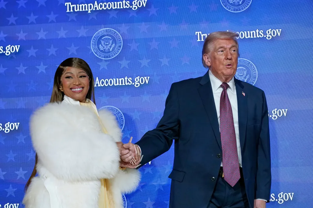 Nicki Minaj and Donald Trump hold hands on stage in front of a blue backdrop with "TrumpAccounts.gov" branding.