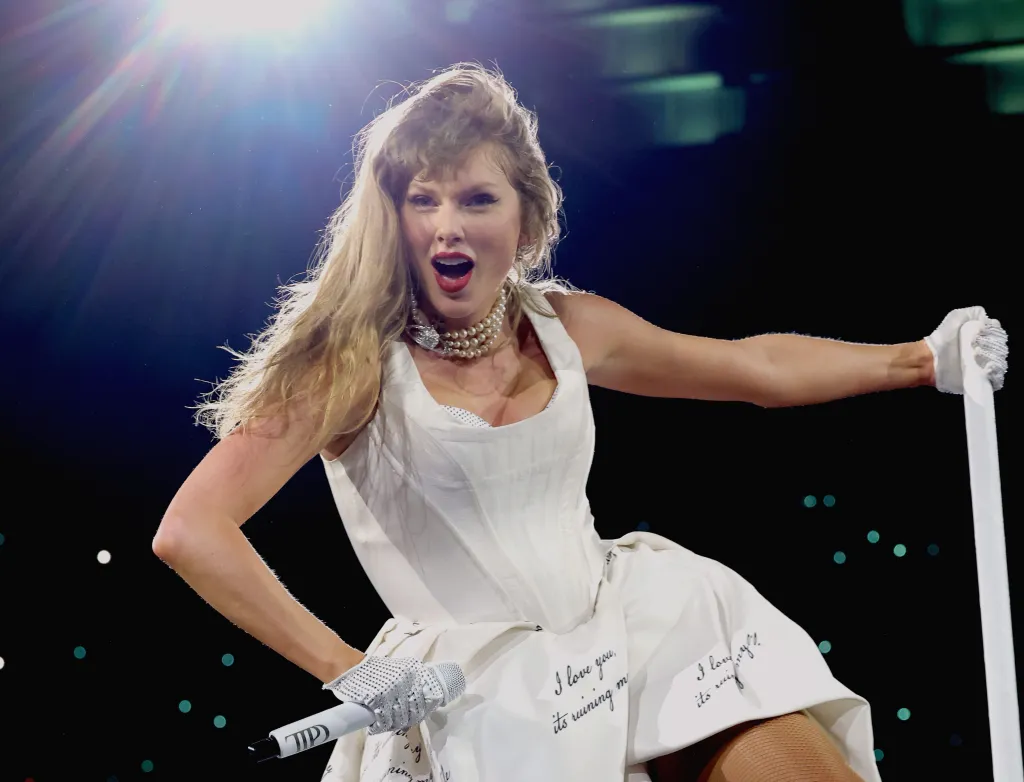 Taylor Swift performing onstage during "The Eras Tour" in Vancouver, wearing a white dress and holding a microphone.