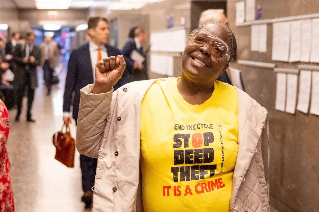 Carmella Charrington in a yellow "STOP DEED THEFT IT IS A CRIME" T-shirt raises her fist in a courthouse.