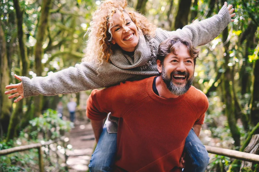 A man carries a woman piggyback as they both laugh in a lush green forest.