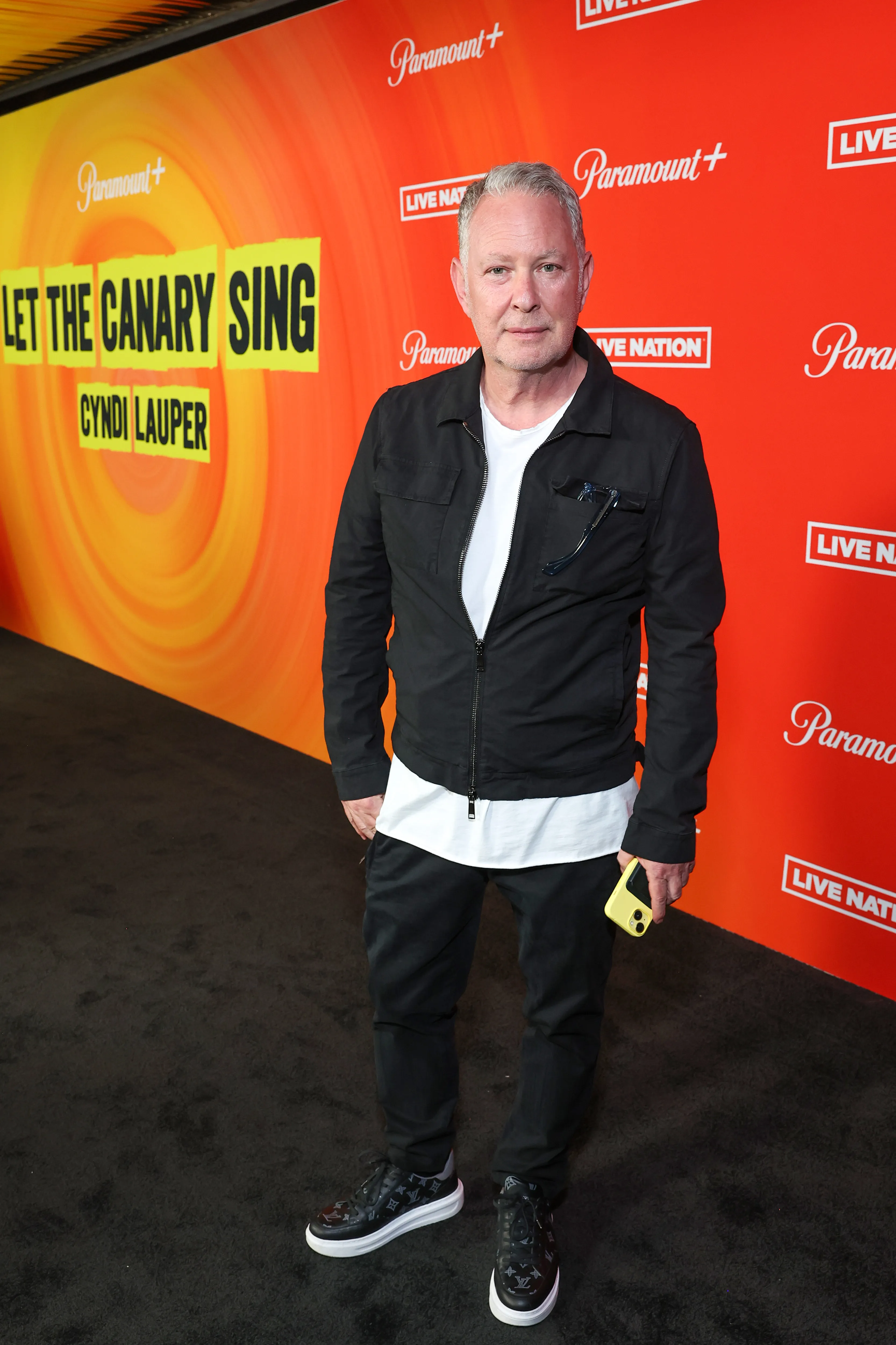Paul Kemsley standing on a black carpet in front of an orange and yellow background with "Let The Canary Sing" and "Cyndi Lauper" visible.