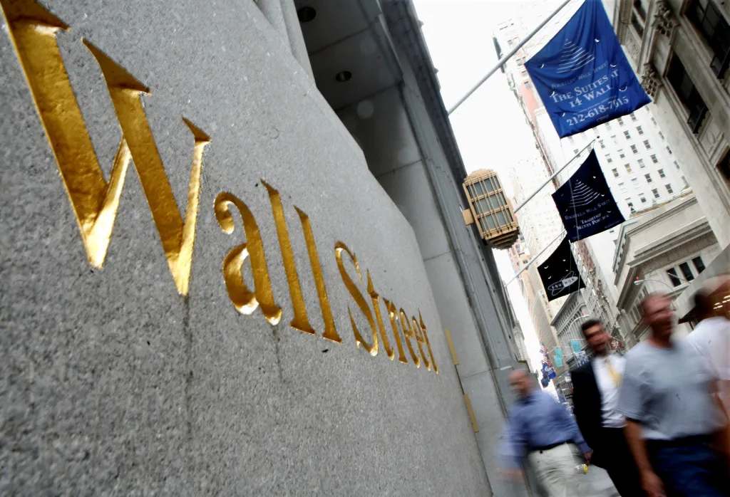 People walk past the gold lettering of "Wall Street" on a building, with flags and other buildings visible in the background.