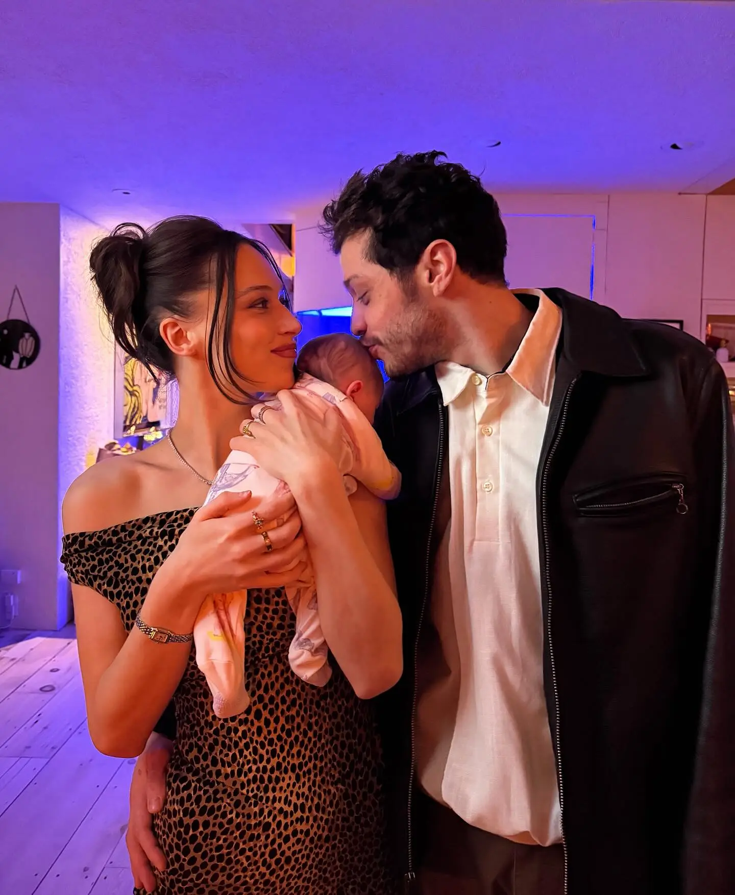 Pete Davidson kissing the forehead of a baby held by a woman in a leopard print dress.