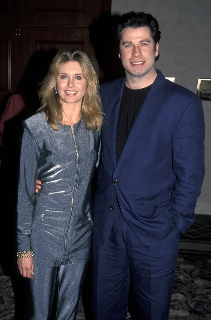 Olivia Newton-John in a silver jumpsuit and John Travolta in a navy blue suit smiling together.