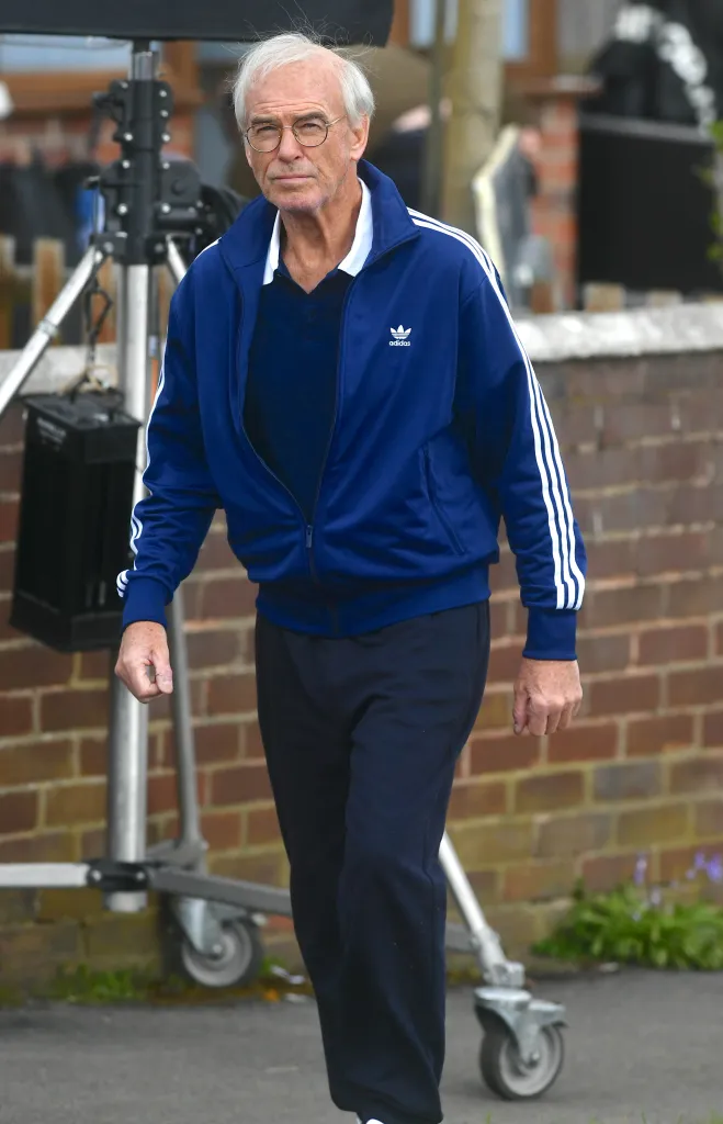 Pierce Brosnan in a blue tracksuit with white stripes, glasses, and grey hair.