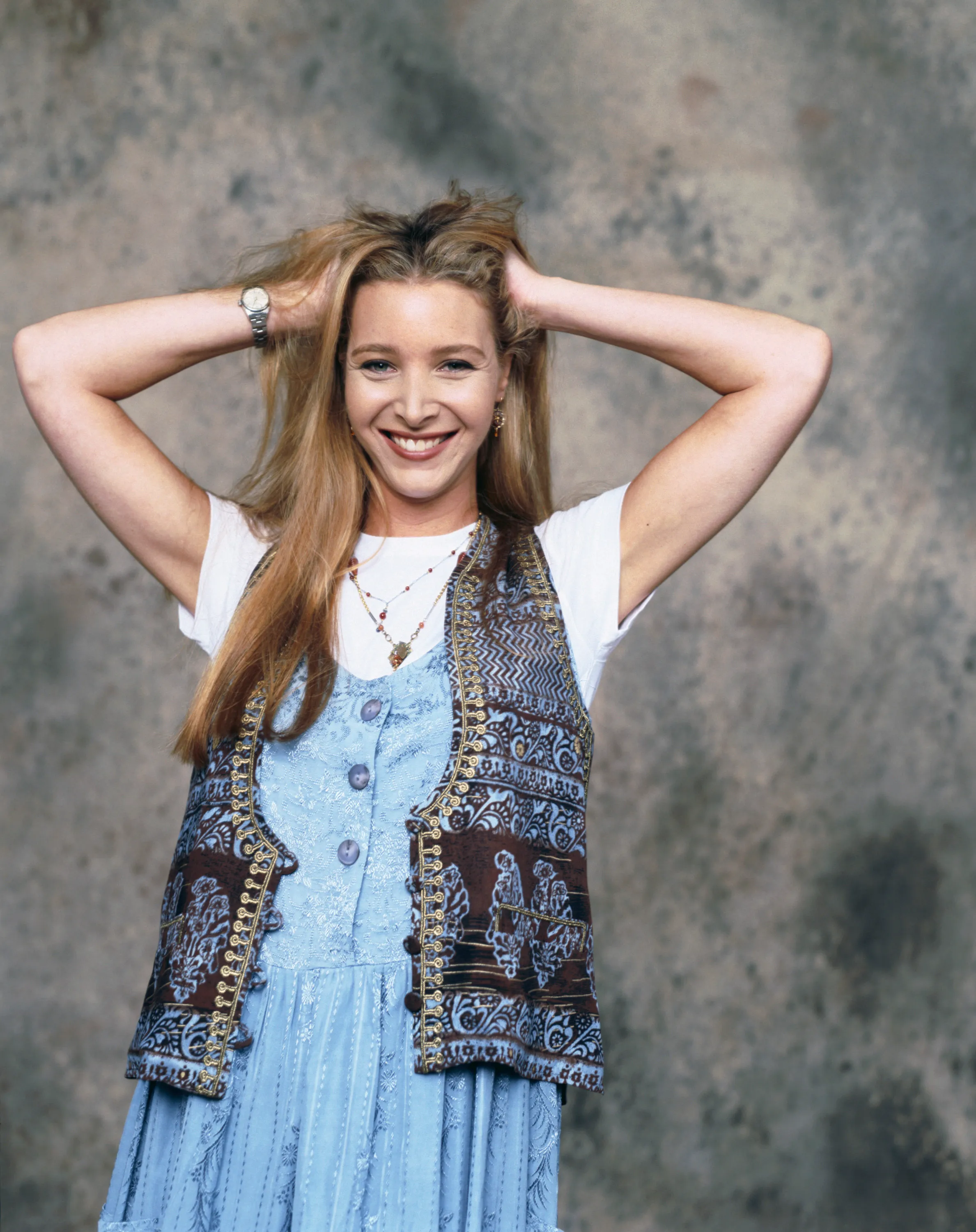 Portrait of actress Lisa Kudrow as Phoebe Buffay in 'Friends' Season 1, smiling at the camera with her hands in her hair, wearing a white t-shirt under a blue floral dress and a brown patterned vest.