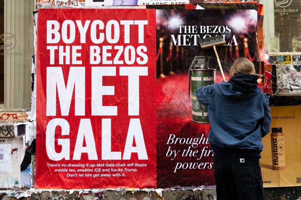 A person with a broom pasting a "Boycott the Bezos Met Gala" poster on a wall in New York City.