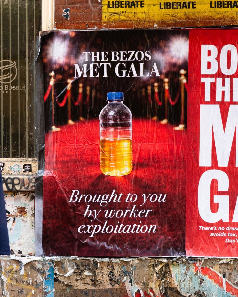 A poster on a wall reads "THE BEZOS MET GALA" with an image of a red carpet and a bottle of yellow liquid. Below, it says, "Brought to you by worker exploitation." Another partial poster to the right reads "BOYCOTT THE MET GALA".