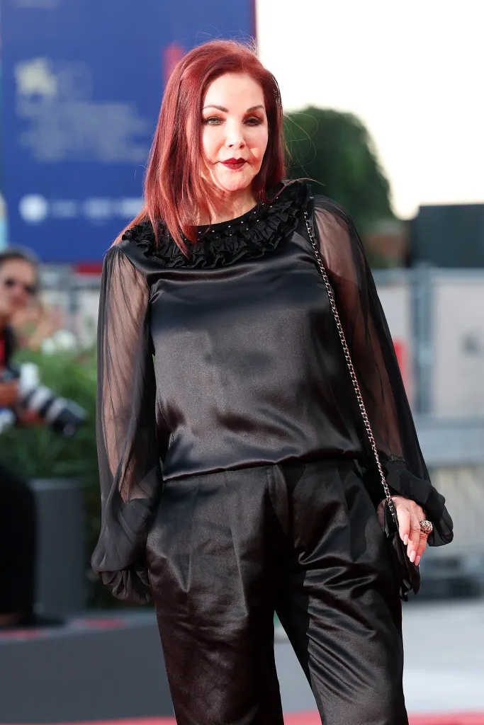 Priscilla Presley walking on a red carpet in a black satin outfit with sheer sleeves.