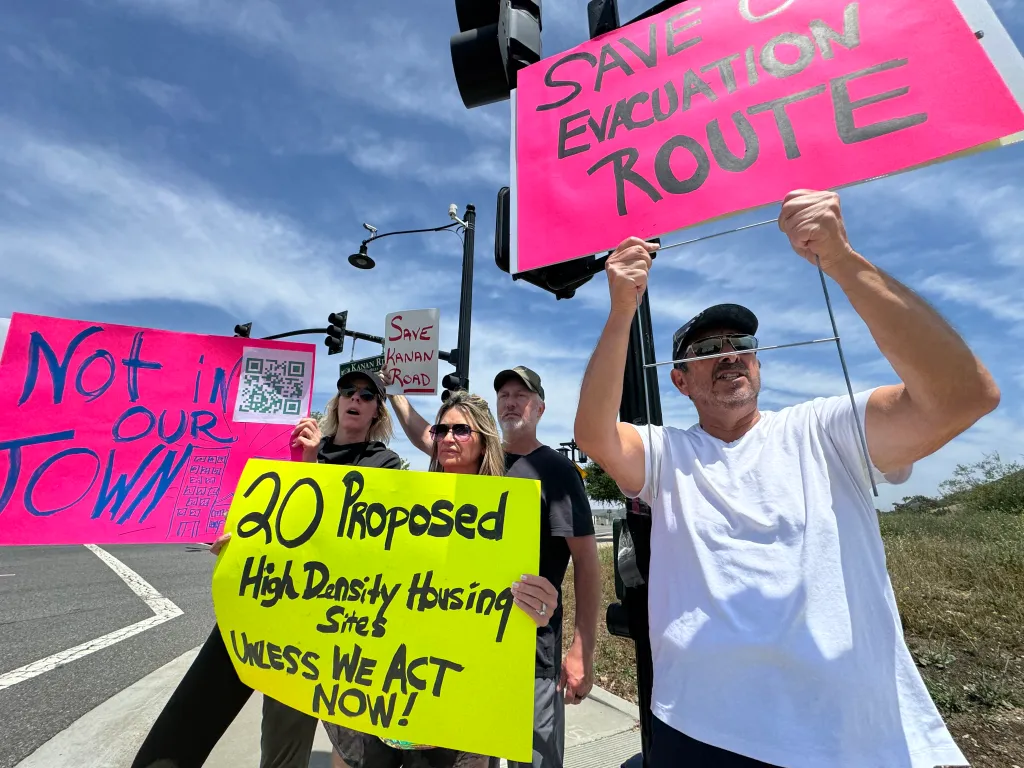 Protesters in Agoura Hills hold signs opposing a state housing plan, with messages including "SAVE EVACUATION ROUTE" and "20 Proposed High Density Housing Sites UNLESS WE ACT NOW!"