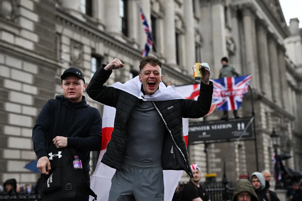 A protester at a "Unite the Kingdom" rally in London wearing an England flag like a cape, pumps his fist and yells, while holding up a canned beverage.