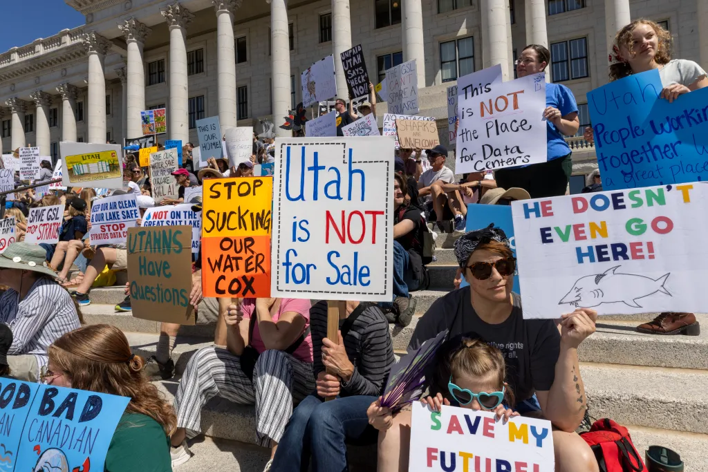 Protesters hold signs against the proposed Stratos data center in Salt Lake City, Utah, with signs such as "Utah is NOT for Sale" and "STOP SUCKING OUR WATER COX."