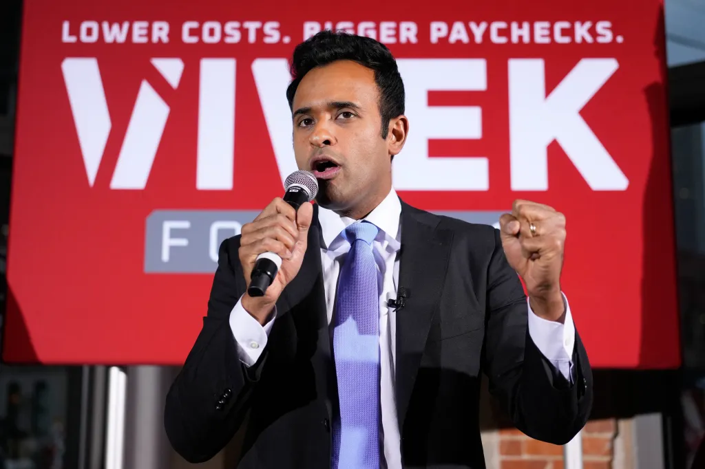 Vivek Ramaswamy speaking into a microphone with a clenched fist, in front of a red banner that reads "LOWER COSTS. BIGGER PAYCHECKS. VIVEK."