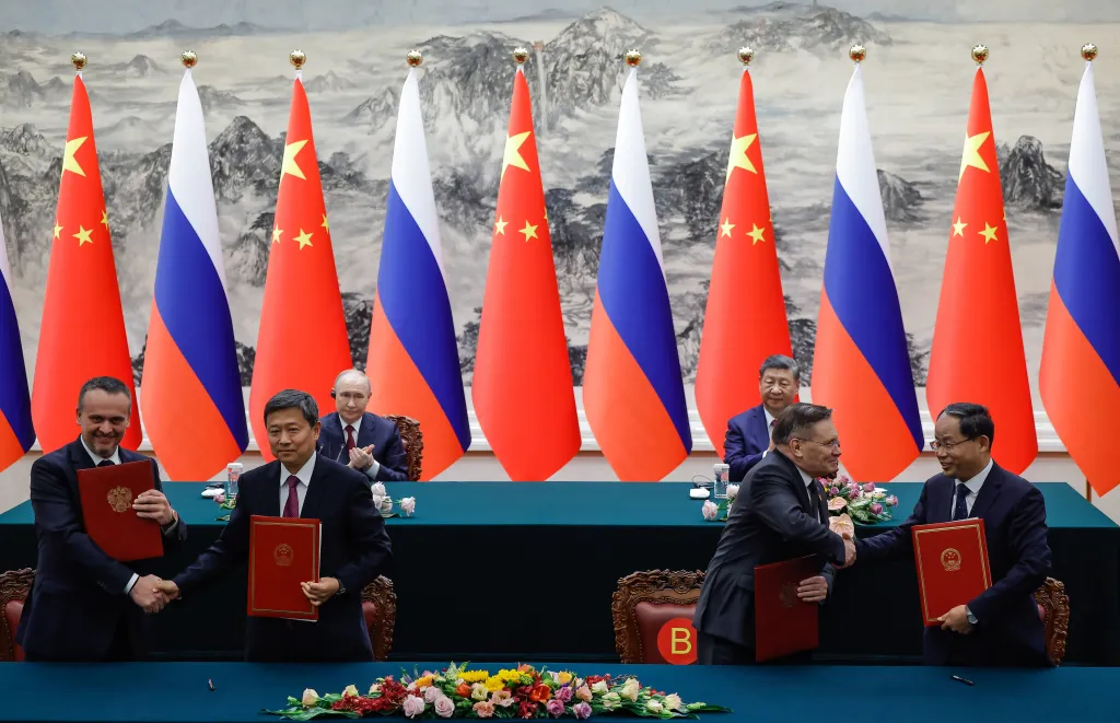 Officials from Russia and China shaking hands after exchanging agreements, with Vladimir Putin and Xi Jinping watching from behind.