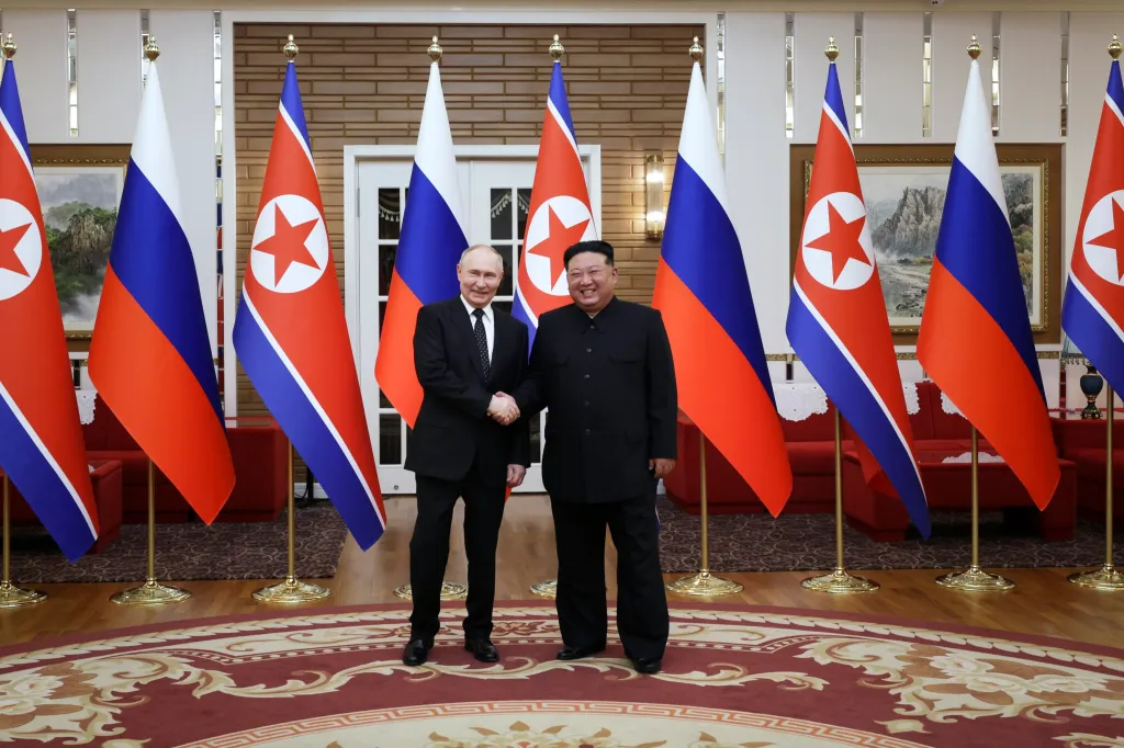 Vladimir Putin and Kim Jong Un shaking hands between North Korean and Russian flags.