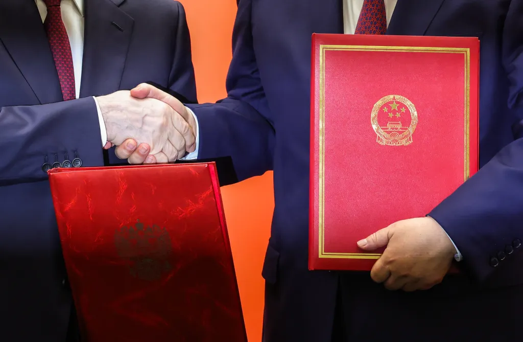 Vladimir Putin and Xi Jinping shake hands while holding agreement folders.