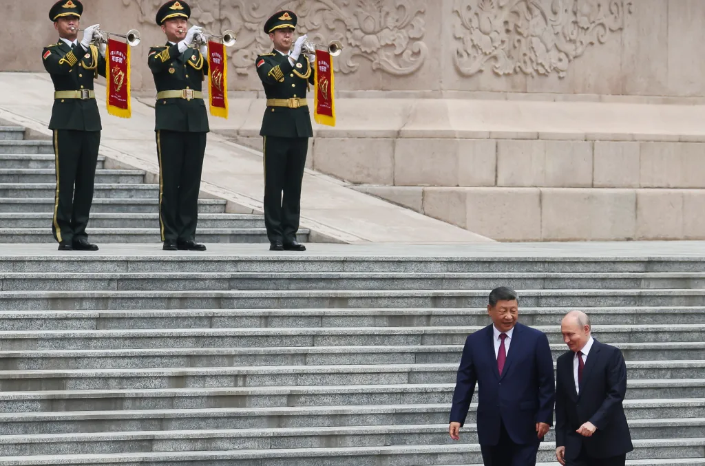 Russian President Vladimir Putin and Chinese President Xi Jinping walk down stairs at a welcoming ceremony, while three soldiers in uniform play trumpets at the top of the stairs.