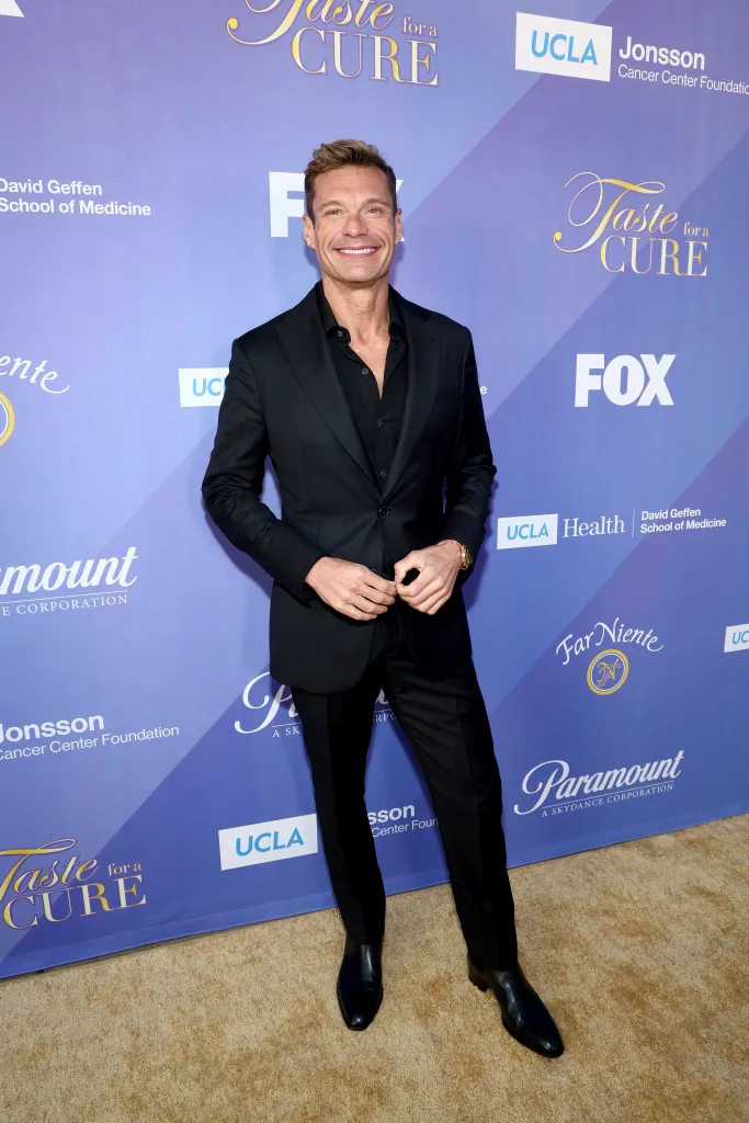 Ryan Seacrest in a black suit with an unbuttoned black shirt smiles at the 29th Annual UCLA Jonsson Cancer Center Foundation's