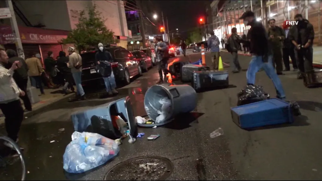 Overturned trash cans and scattered litter on a wet street at night with people and cars in the background during a protest.