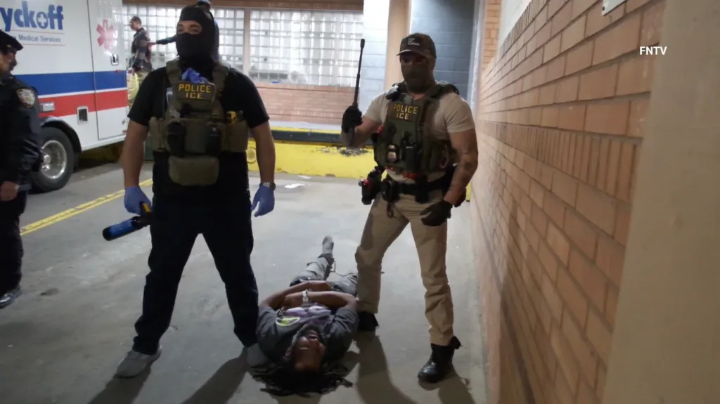 Two ICE officers stand over a man lying on the ground, with a Wyckoff Medical Services ambulance in the background.