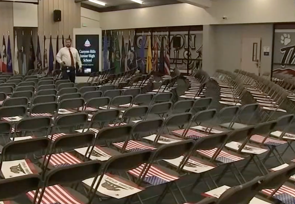 An empty school auditorium set up for an assembly with rows of chairs, each featuring a small US flag and a California Republic flag on the seat.