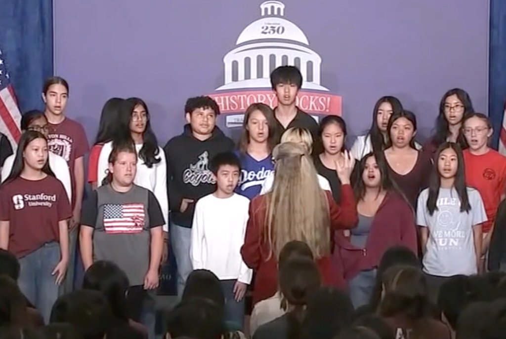 A group of middle and high school students sings on stage in front of a banner that reads "History Rocks" and an American flag.