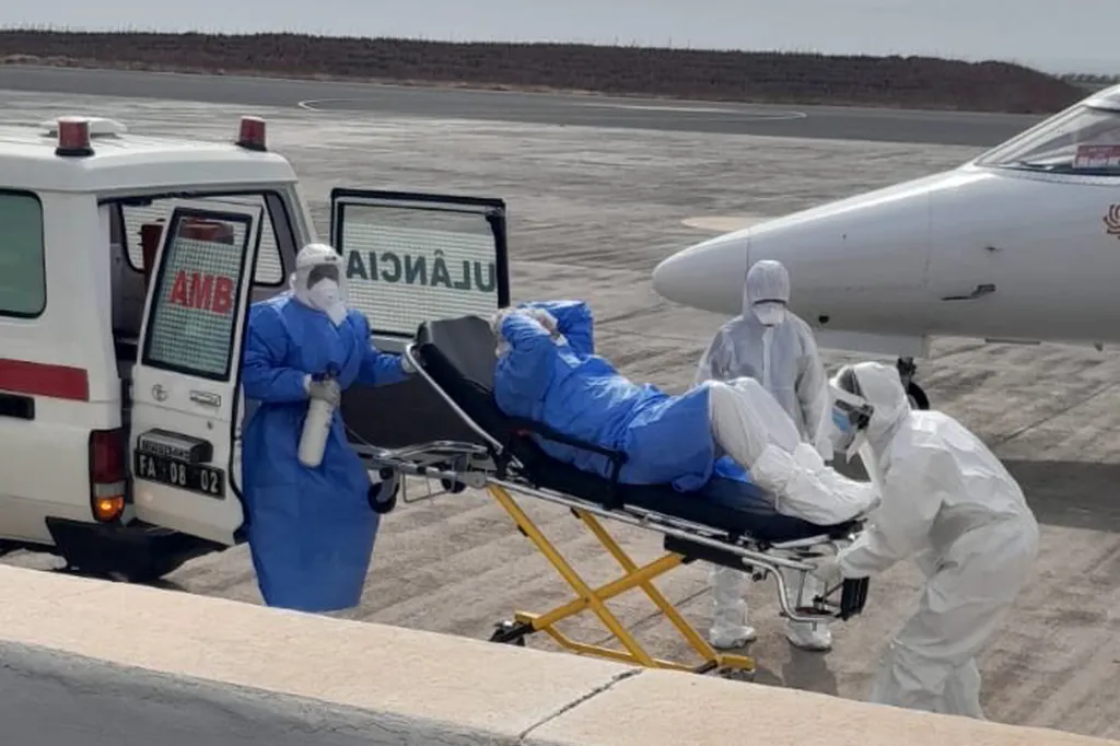 Medical personnel in protective suits move a patient on a stretcher from an ambulance towards an aircraft.