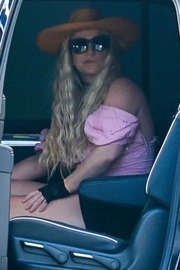 Britney Spears in a wide-brim hat, sunglasses, pink off-shoulder top, black shorts, and black lace gloves sits in a car after returning from Cabo.