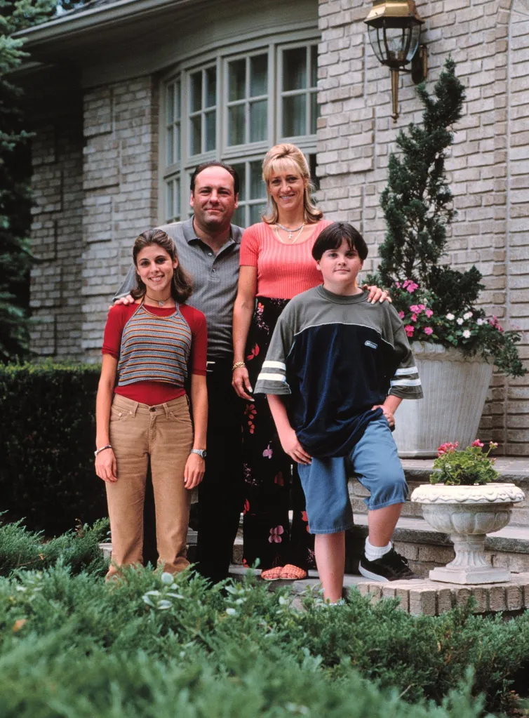 Jamie Lynn Sigler, James Gandolfini, Edie Falco, and Robert Iler from The Sopranos posing in front of a house.