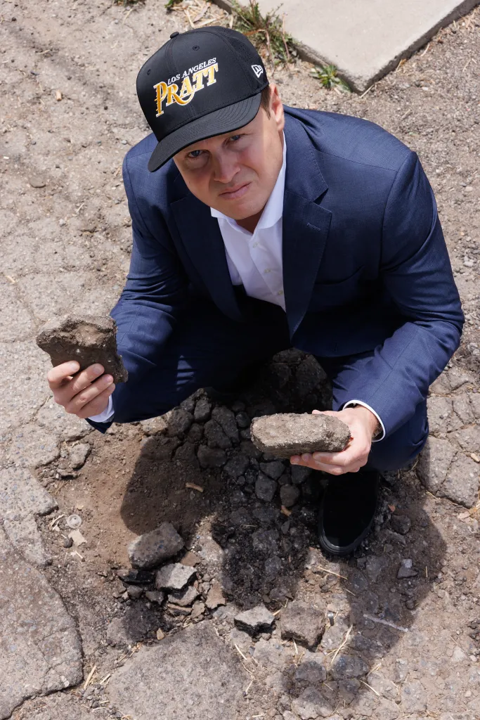 Spencer Pratt, in a suit and a "Los Angeles Pratt" hat, kneels next to a pothole, holding pieces of asphalt.