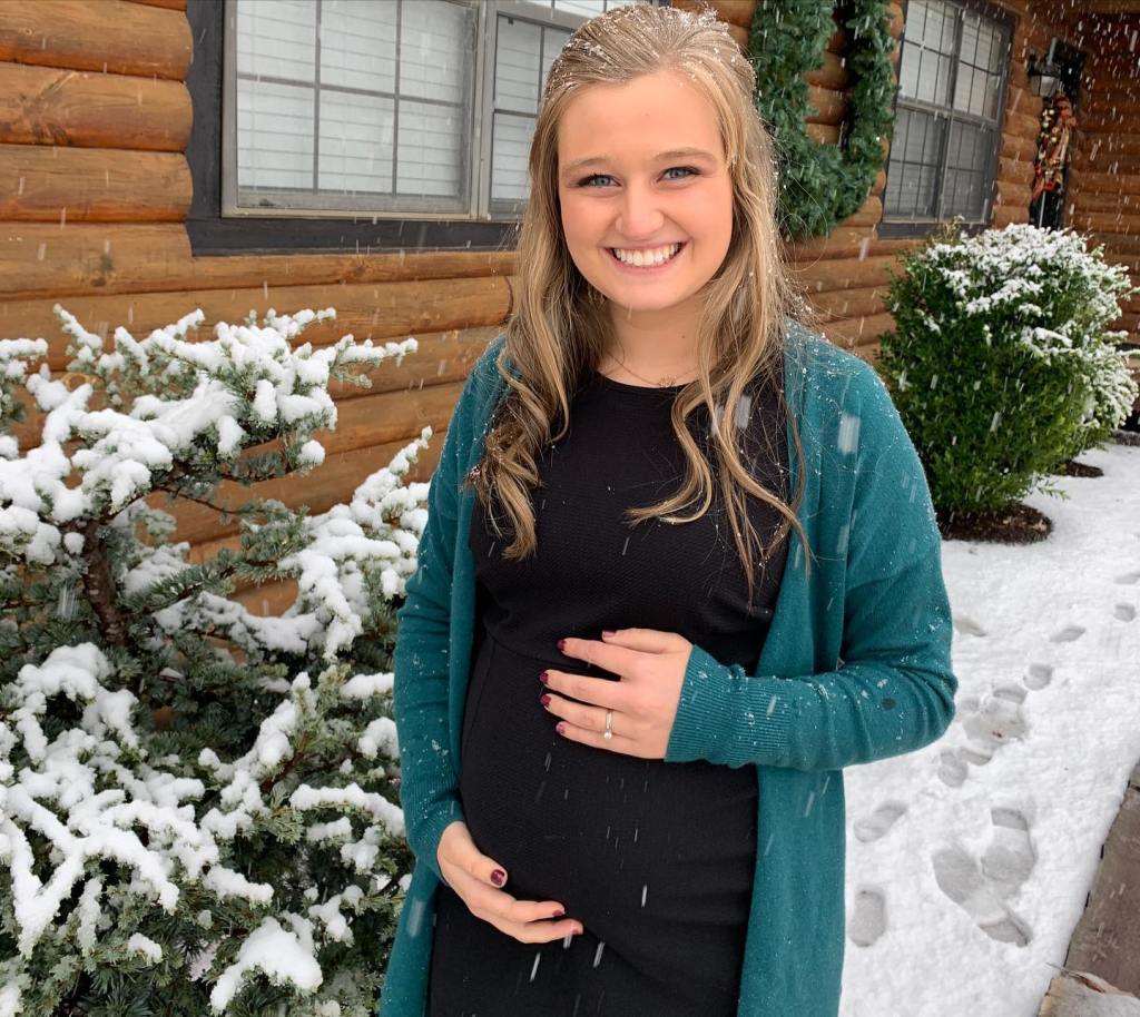 Pregnant woman in a black dress and teal cardigan, smiling outdoors in the snow.