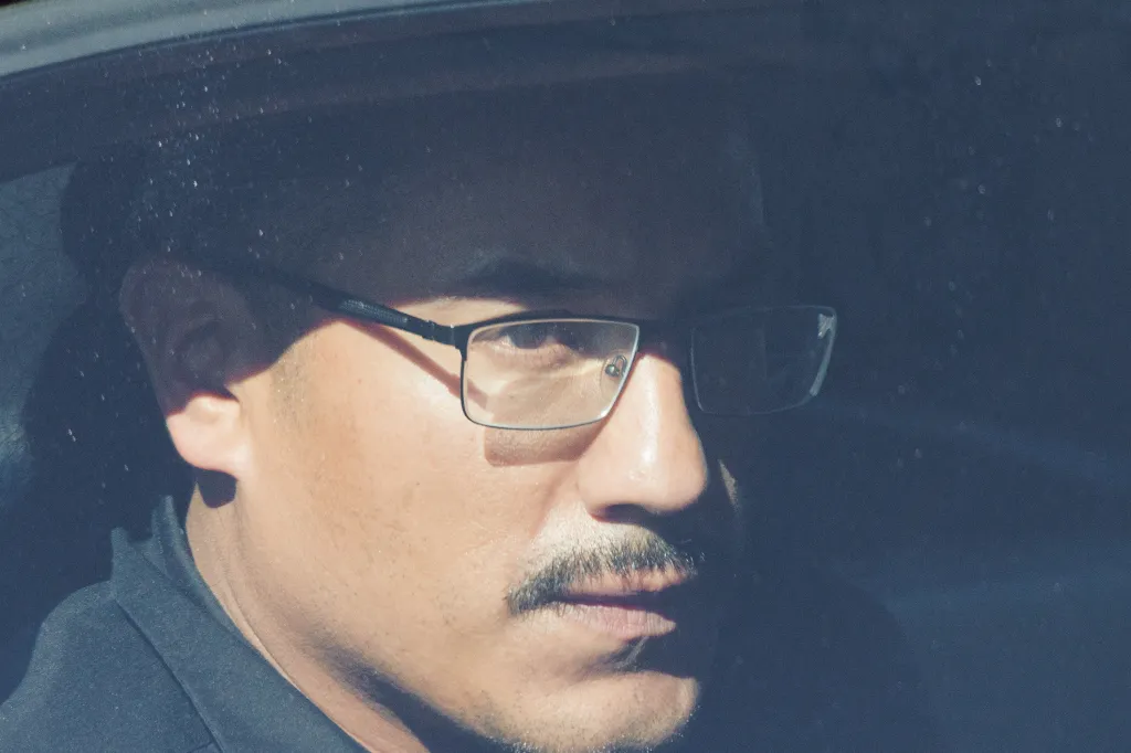 Close-up of a suspect's face, wearing glasses, partially obscured by shadows and reflections on a window.