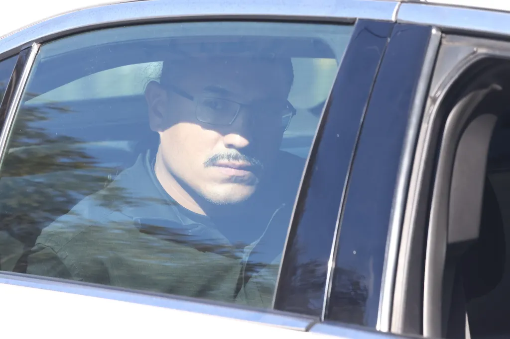 A suspect in glasses and a mustache looks out the window of a car.