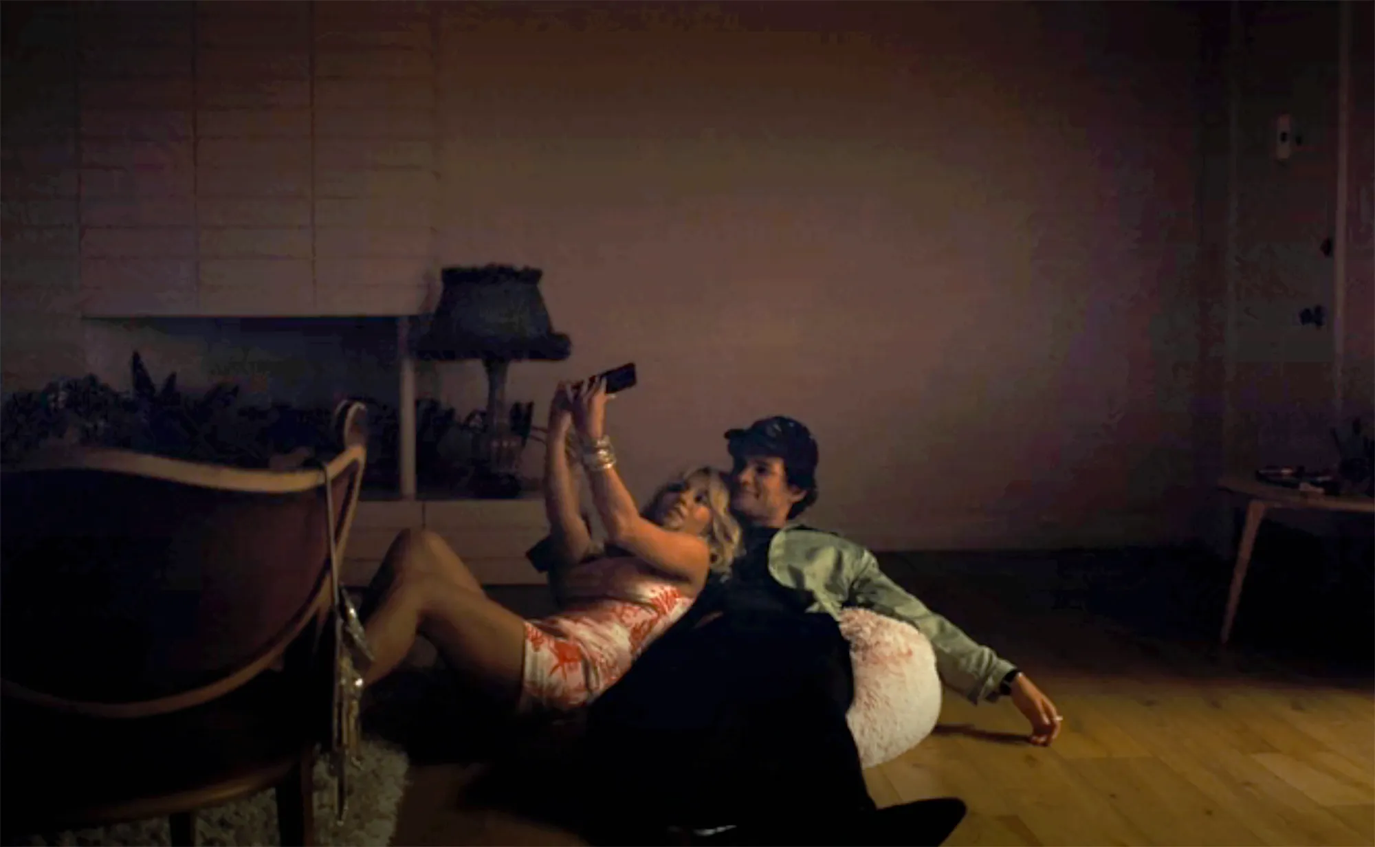 Sydney Sweeney in a floral dress takes a selfie with a Homer Gere in a black cap and jacket, both lying on a wooden floor in a dimly lit room.