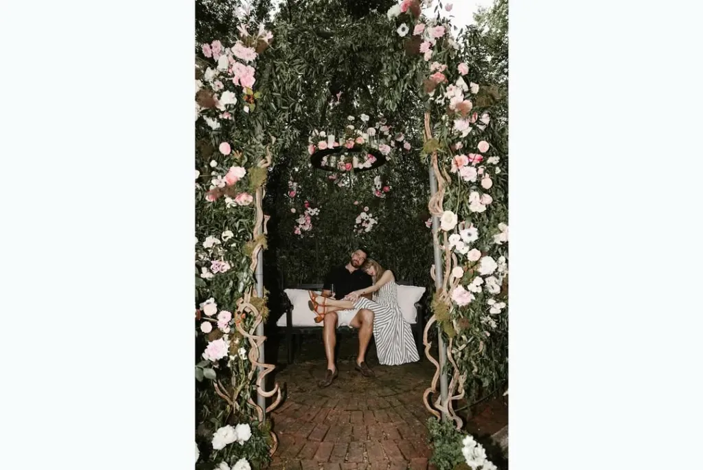 Taylor Swift and Travis Kelce seated on a white couch under a floral archway.