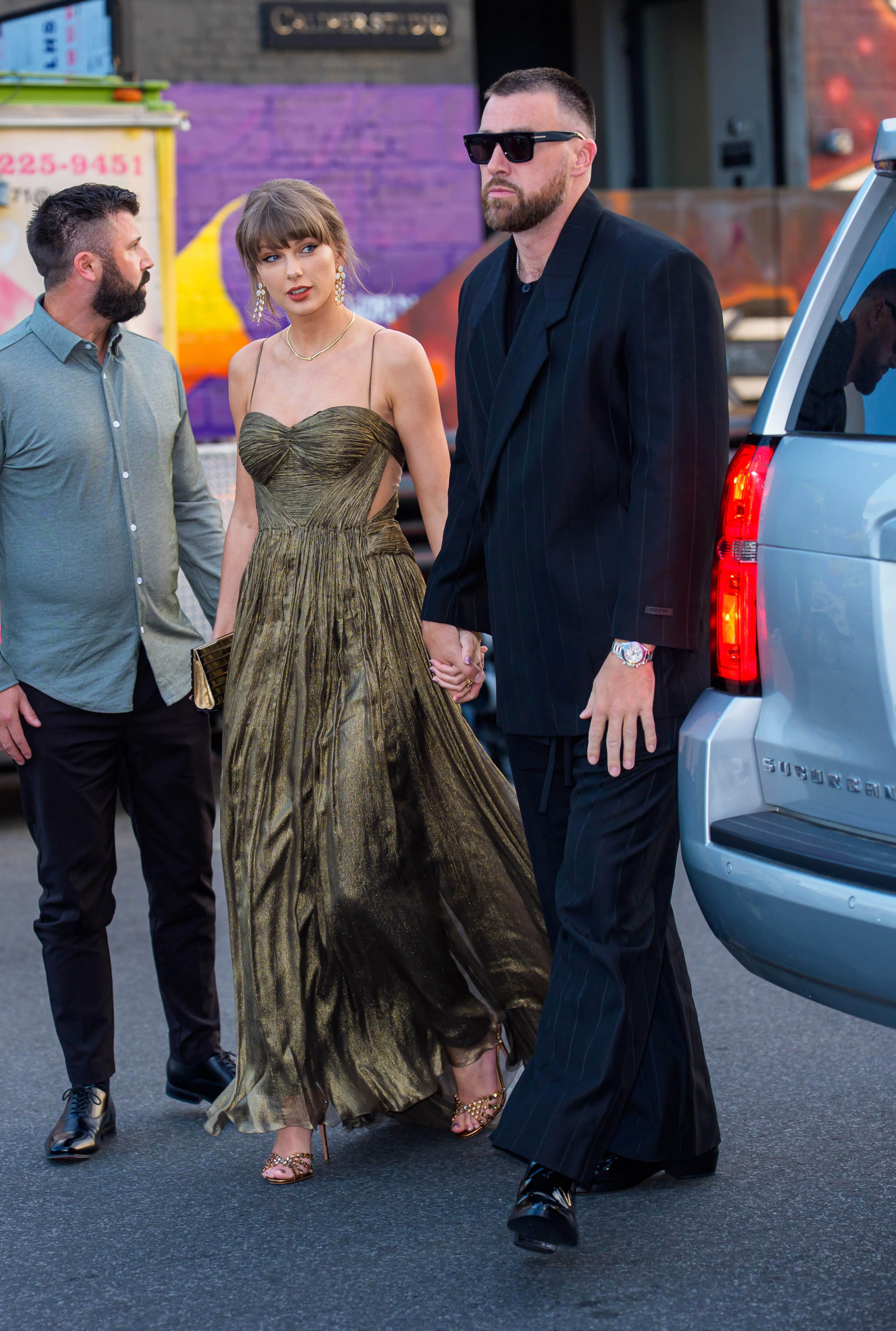 Taylor Swift in a gold dress and Travis Kelce in a dark suit holding hands.