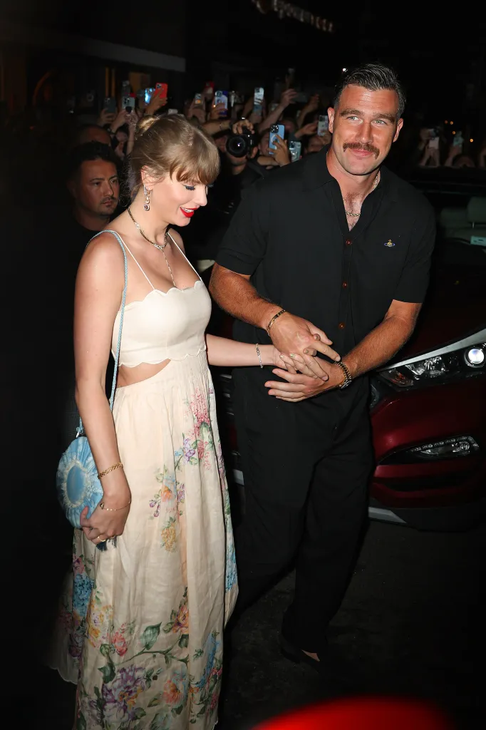 Taylor Swift and Travis Kelce holding hands while walking, with onlookers in the background.