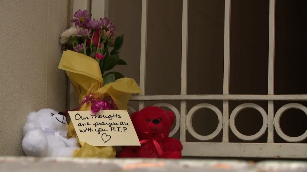 A memorial with flowers, two teddy bears, and a handwritten note that reads, "Our thoughts and prayers are with you. R.I.P."