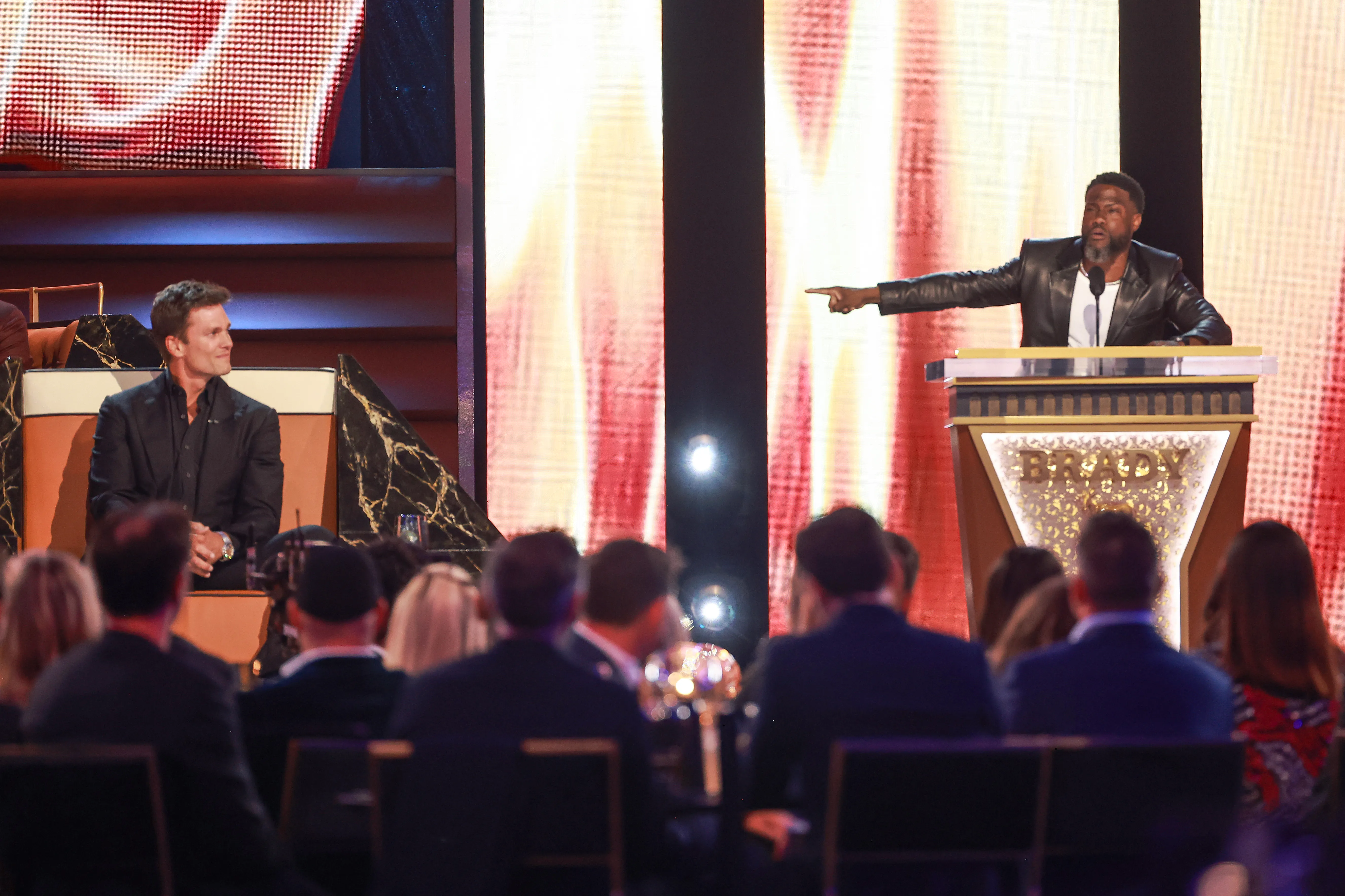 Kevin Hart speaking at a podium and pointing, with Tom Brady sitting and smiling in the background.
