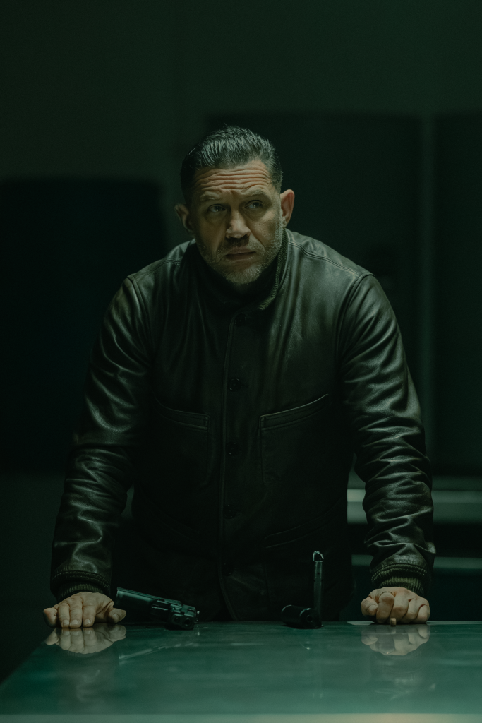 Tom Hardy as Harry Da Souza in MOBLAND, wearing a dark leather jacket, standing at a table with a gun and a gun magazine.