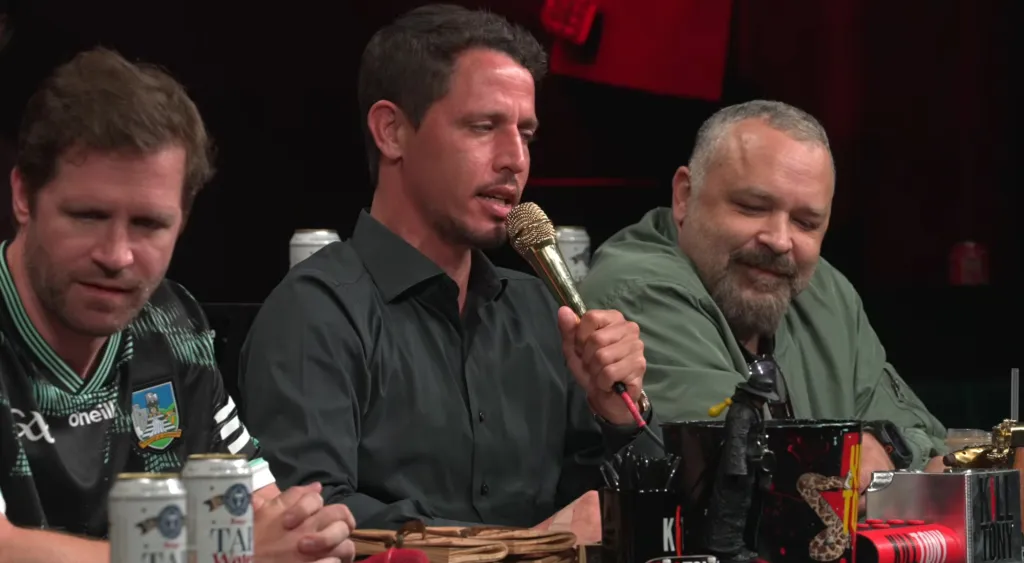 Tony Hinchcliffe holding a golden microphone, sitting between two other men.
