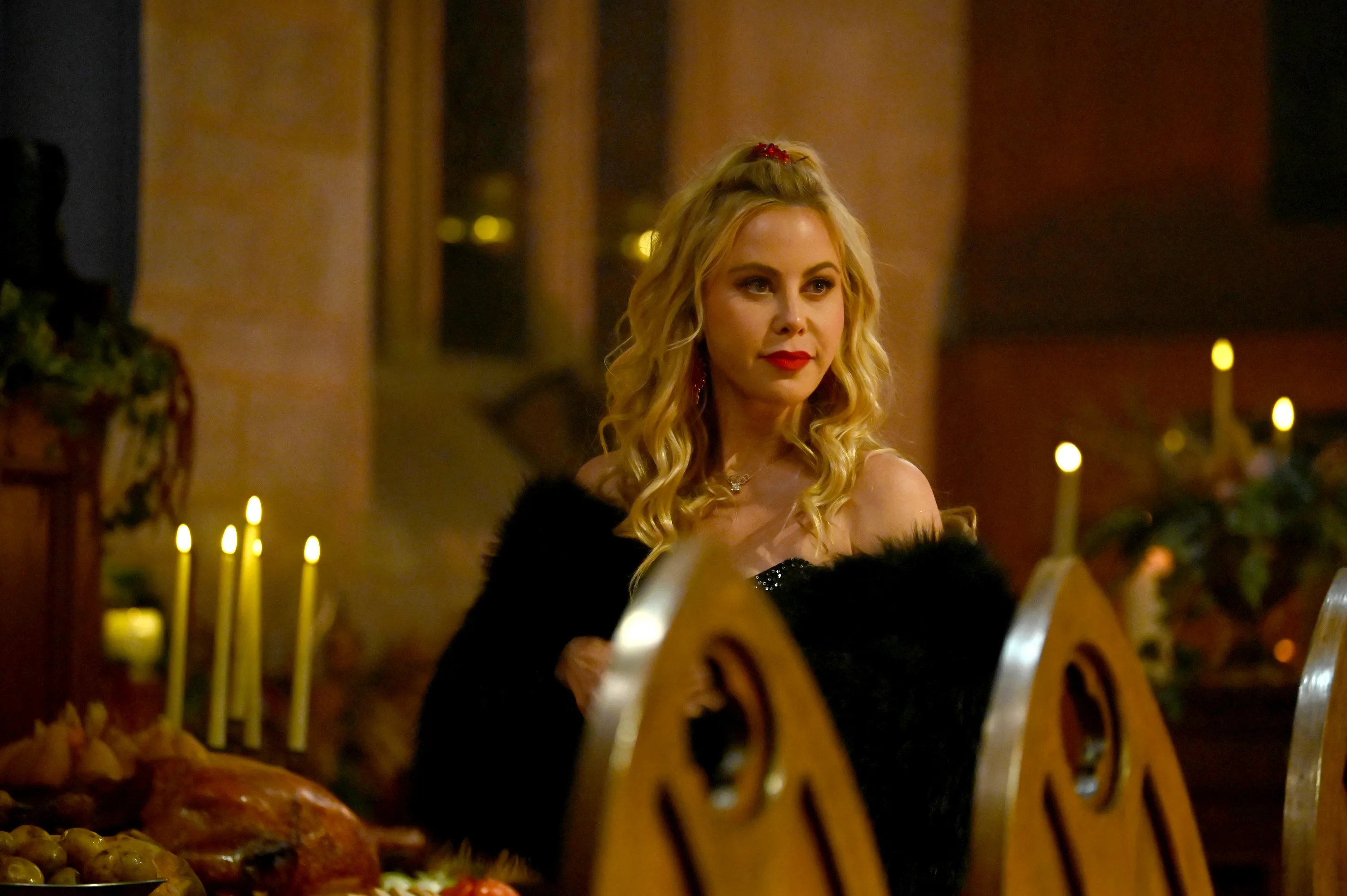 Tara Lipinski with long blonde hair and a dark feathered top, red lipstick and earrings, in a banquet setting with candles and food on the table.